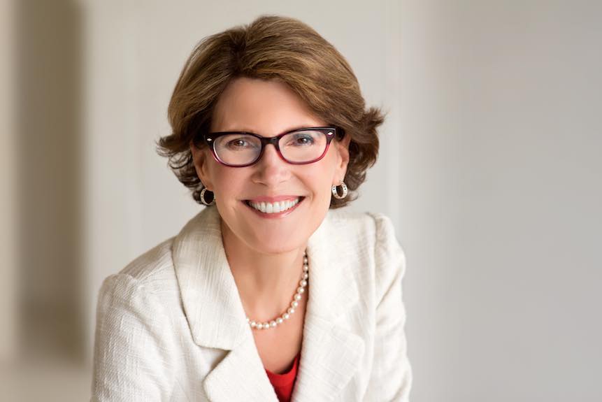 Shows a professional business headshot of a woman in white jacket, pearls, glasses, smiling