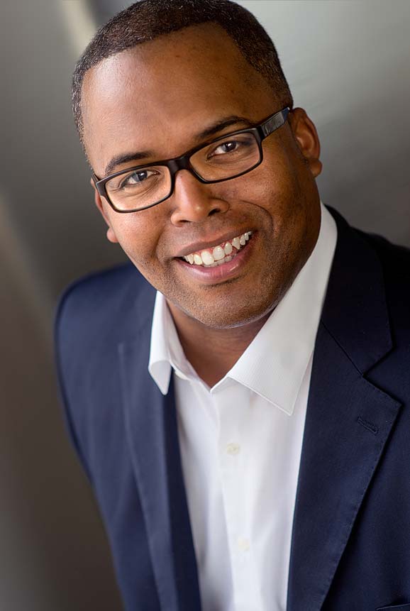 Shows a professional business headshot of a man in blue jacket, glasses, smiling