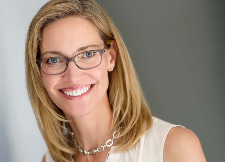 Shows a professional business headshot of a woman waering chain necklace, glasses, smiling