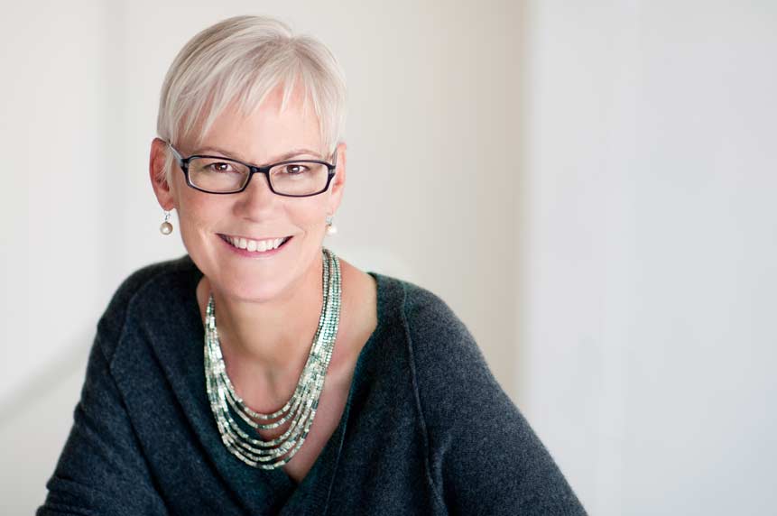 Shows a professional business headshot of a casually, but well-dressed woman wearing glasses and pearl earrings